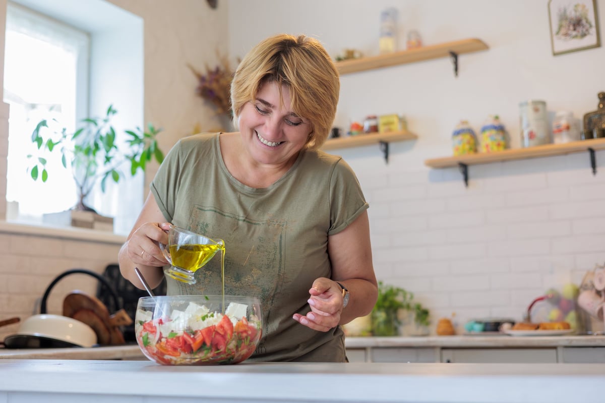 Smiling mature woman enjoys preparing a healthy salad with fresh tomatoes and greens in her kitchen, adding spices. Her happy expression and focus highlight self-care, balanced nutrition, and mindful wellness relevant to women's health and hormonal balance.