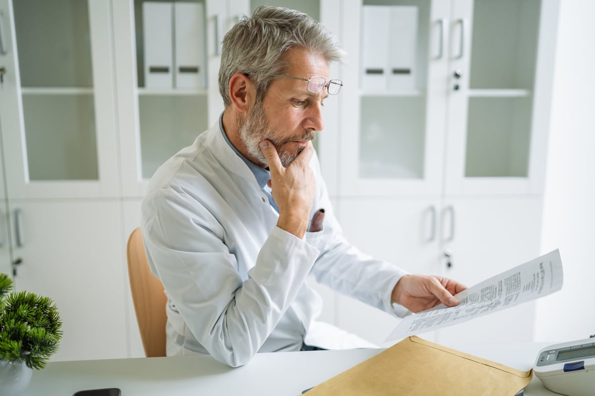 Experienced senior male doctor reviewing medical documents with a thoughtful expression, analyzing patient information in a modern clinic setting, demonstrating professional expertise