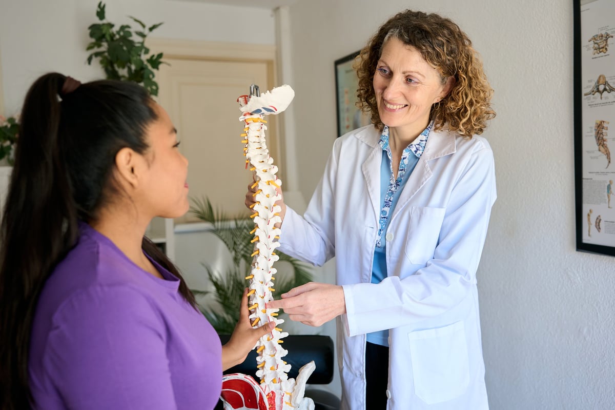 Doctor in lab coat educating a female patient about human spine anatomy using a skeletal model during an appointment